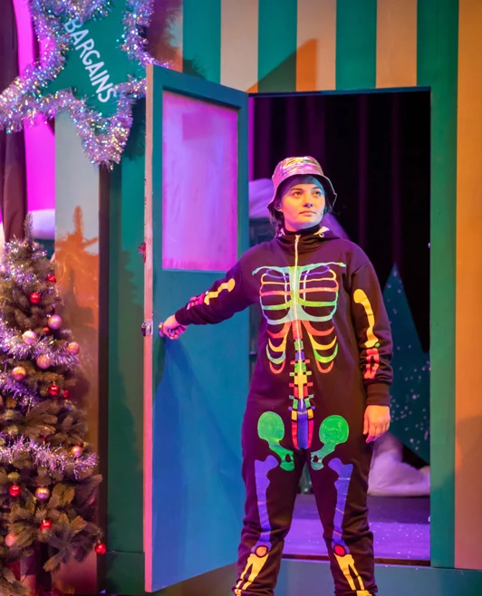 Performer dressed in a colorful skeleton costume, opening a door to a whimsical, festive set. Performer dressed in a colorful skeleton costume, opening a door to a whimsical, festive set.